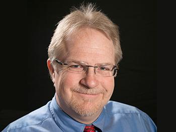 Co-Director wearing glasses and a blue collared shirt with red tie smiling for the camera