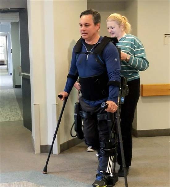 Male research participant with spinal cord injury wears and walks in Ekso GT assisted by female physical therapist standing in back of him