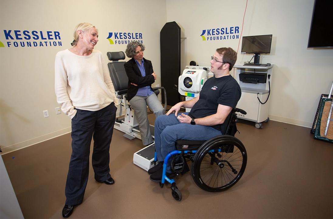 Three individuals in a rehabilitation research lab at Kessler Foundation, including one person using a wheelchair, engaged in discussion near advanced rehabilitation equipment and monitors. 