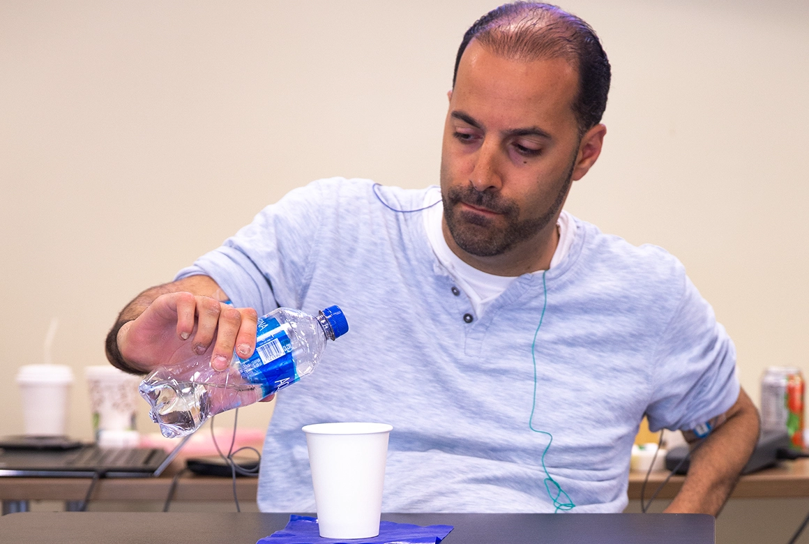 Individual seated at a table pouring water from a plastic bottle into a paper cup during a rehabilitation or therapy activity, wearing a light gray shirt with wires attached for monitoring. 