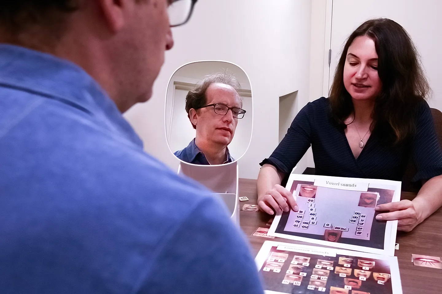 A man wearing glasses viewing himself in a mirror while practicing reading after stroke with a female clinician sits on opposite side of table.