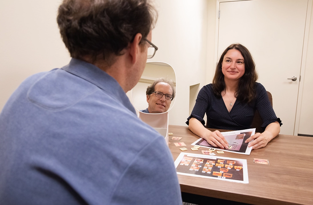 Two individuals seated at a wooden table during a therapy or rehabilitation session, using printed picture cards and a mirror as part of a communication exercise. 