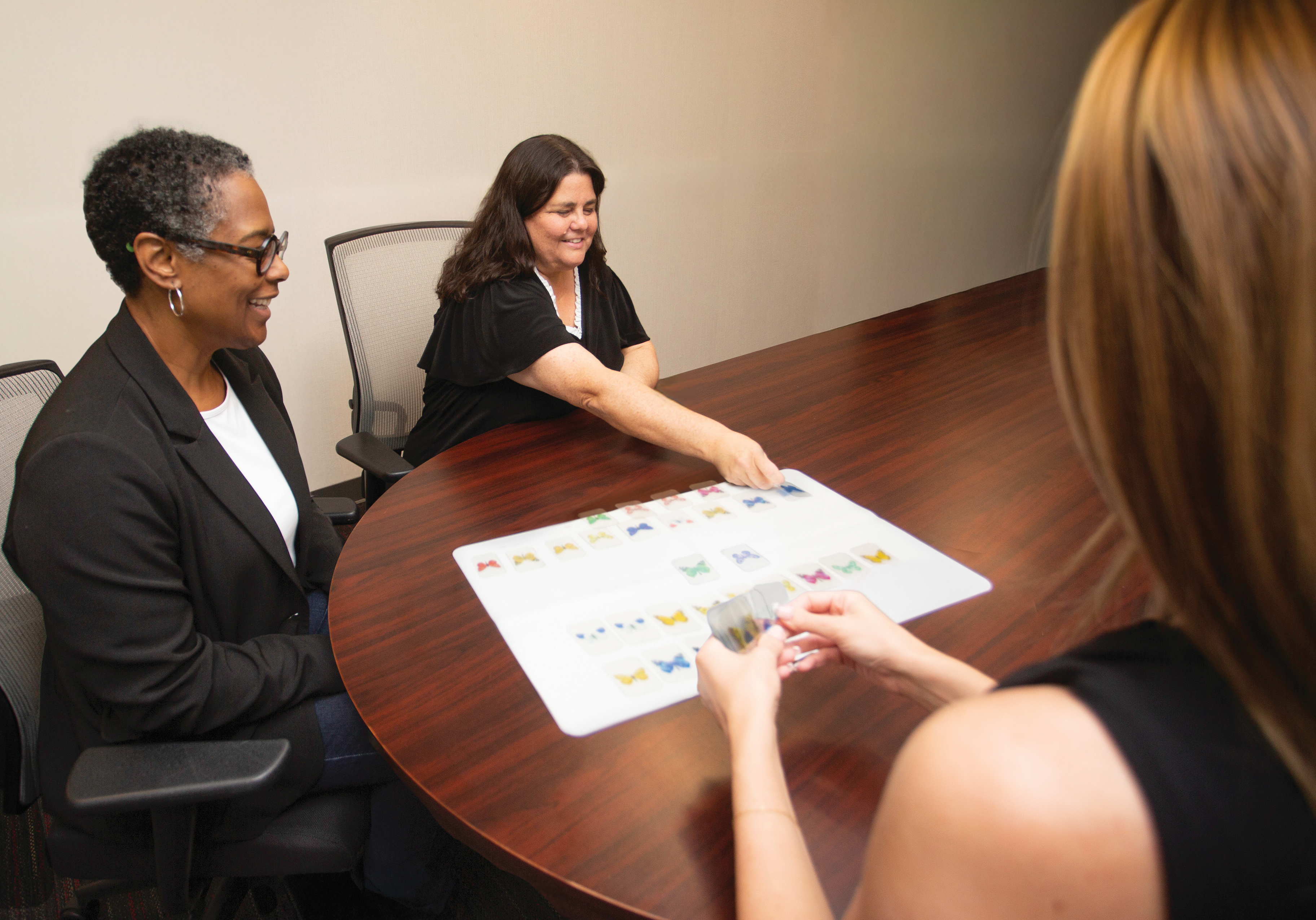 Three individuals are seated around a large wooden conference table engaged in an activity. A laminated sheet with colorful symbols or images is placed on the table, and one person is pointing to it. Another person is holding a stack of cards, suggesting a cognitive or language-based exercise in a professional or therapeutic setting.