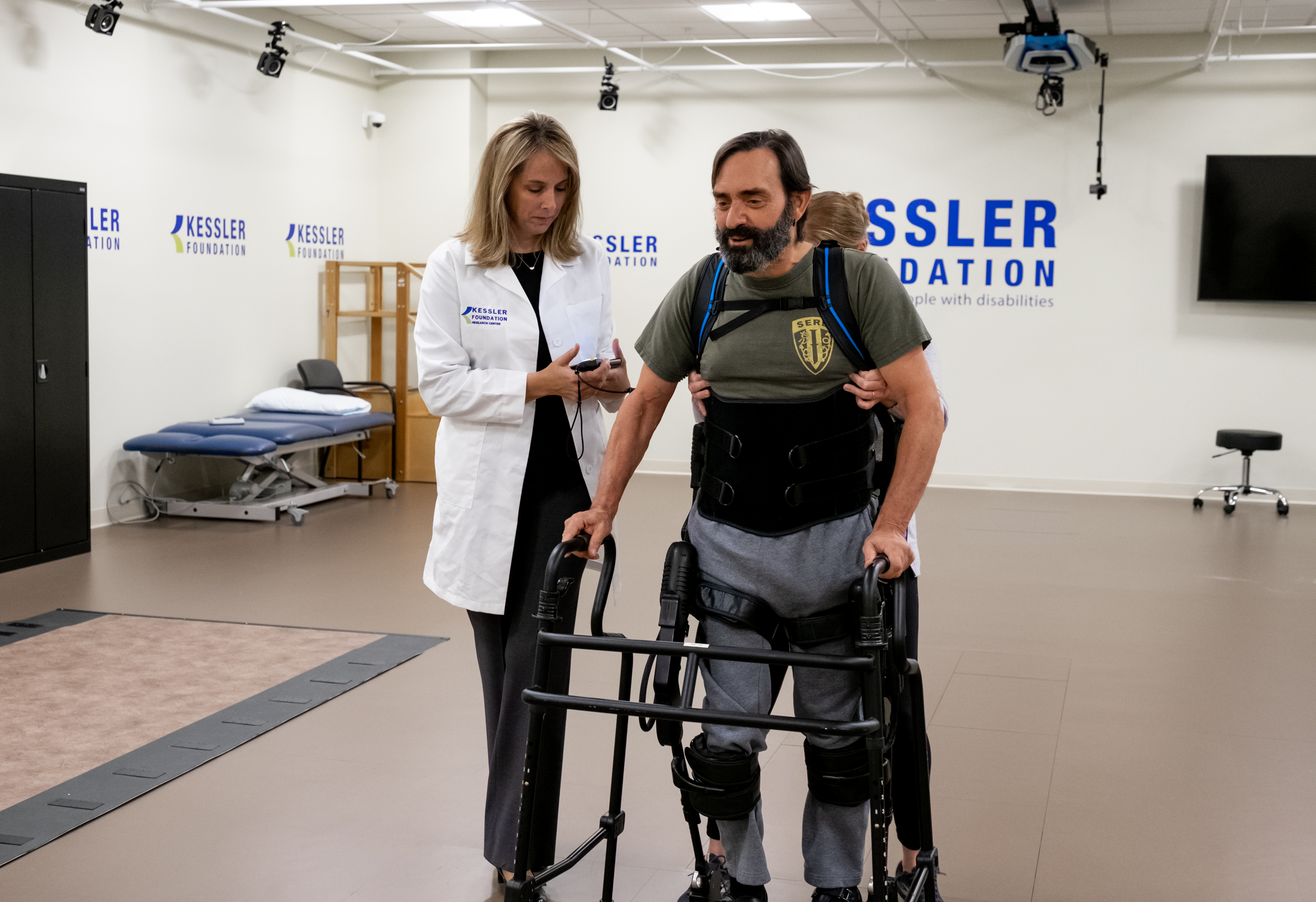 A rehabilitation training session in a clinical setting. A person wearing a robotic exoskeleton is using a walker for support while another individual assists from behind. A clinician in a white lab coat stands nearby holding a tablet. The background includes a treatment table, exercise equipment, and a wall with the Kessler Foundation logo.