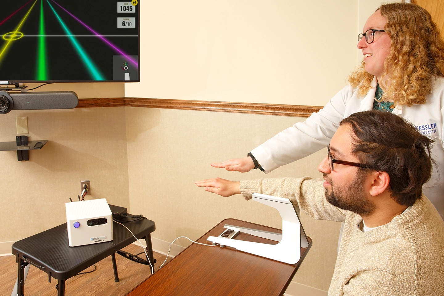 Female clinician shows male stroke participant performing how to perform rehabilitative hand exercises while sitting in front of a screen that shares a gaming element to make the exercises more interactive.