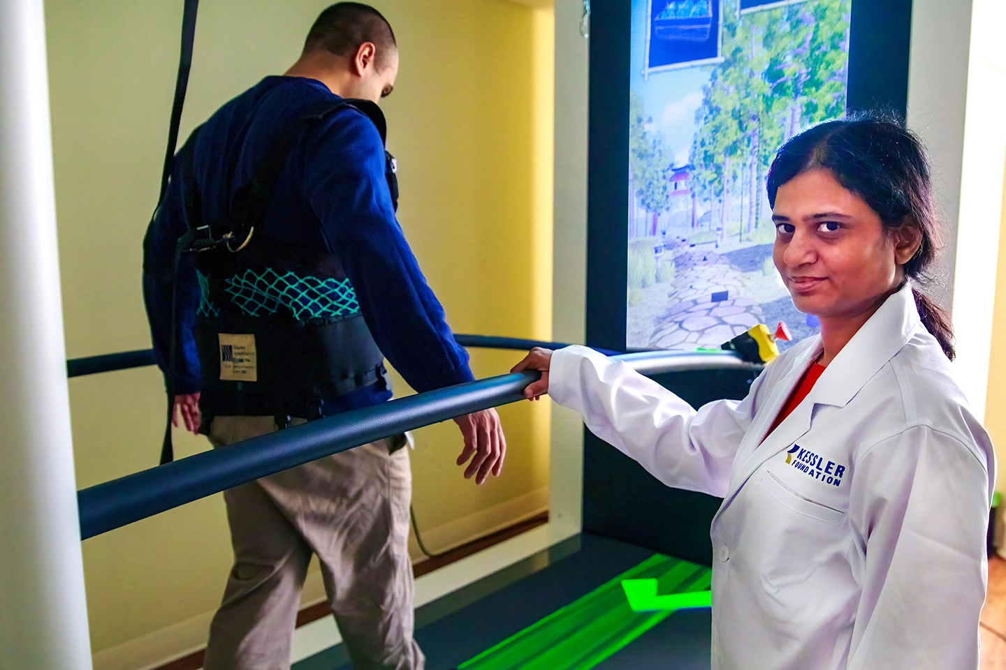 Person wearing a white lab coat standing beside a treadmill, monitoring another individual walking with a safety harness and support bars, in a rehabilitation setting with a virtual reality screen in the background.