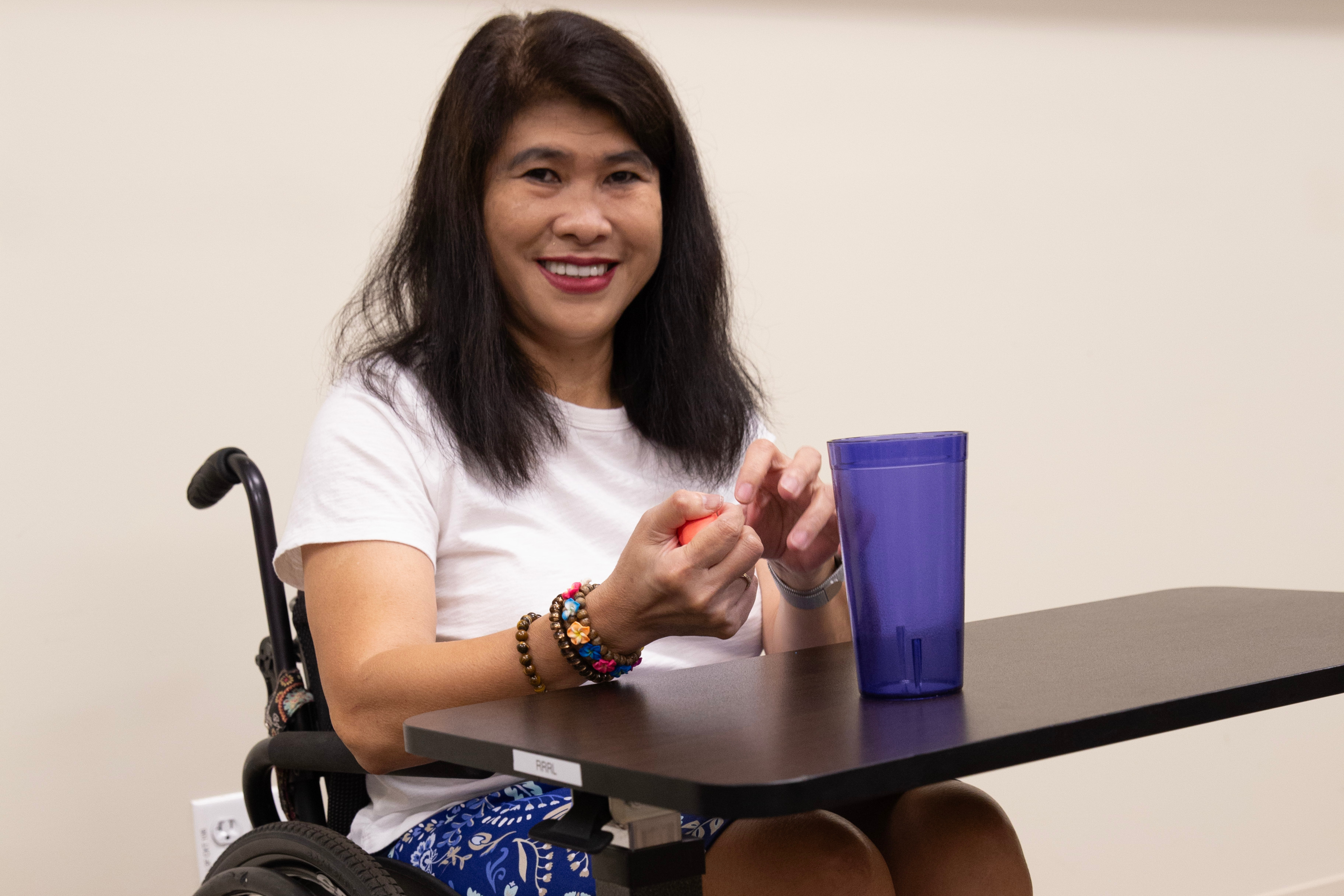A person seated in a green-framed wheelchair at a small black table, wearing a white short-sleeve shirt and blue patterned shorts. The individual is holding an object in their hands, and a large translucent purple cup is placed on the table. The background features a beige wall with the Kessler Foundation logo repeated across the top.