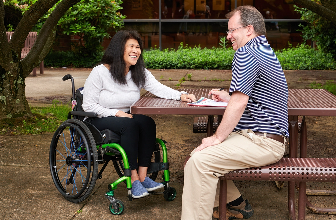 Accessible outdoor setting with a person in a wheelchair and another individual reviewing papers at a picnic table. 