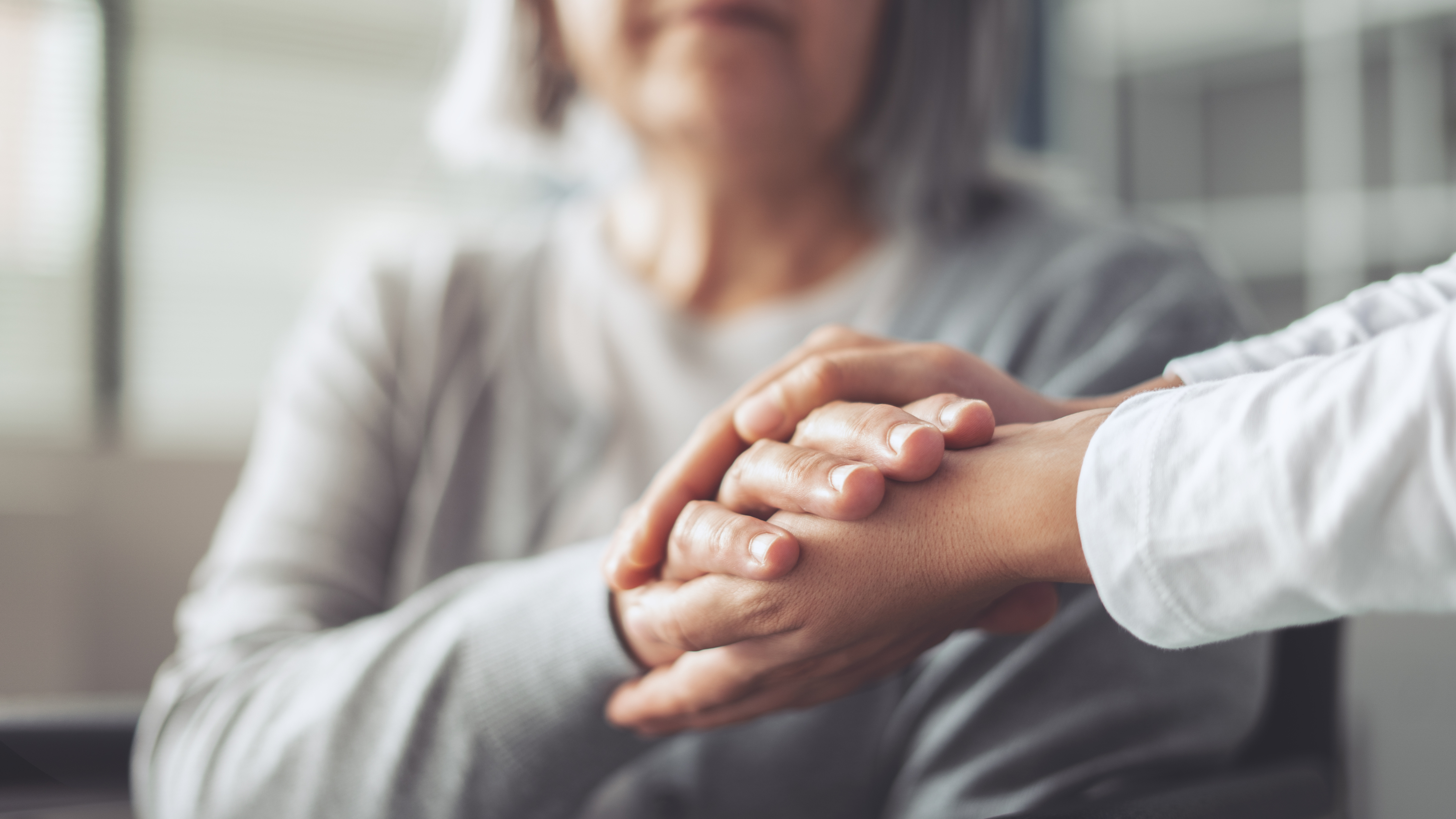 Close-up of two people’s hands in a comforting gesture. One person is gently holding the other’s hand with both hands. The background is softly blurred, showing part of the upper body of one individual wearing a light gray top. The image conveys support, care, and connection.