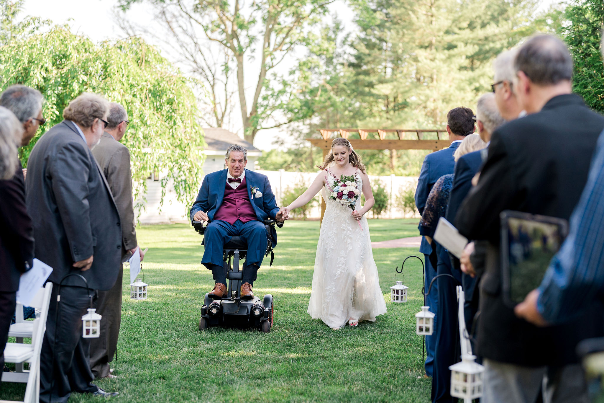 A father using a power wheelchair escorts his daughter down the aisle at her outdoor wedding, as guests stand on either side in a garden setting surrounded by trees and greenery.