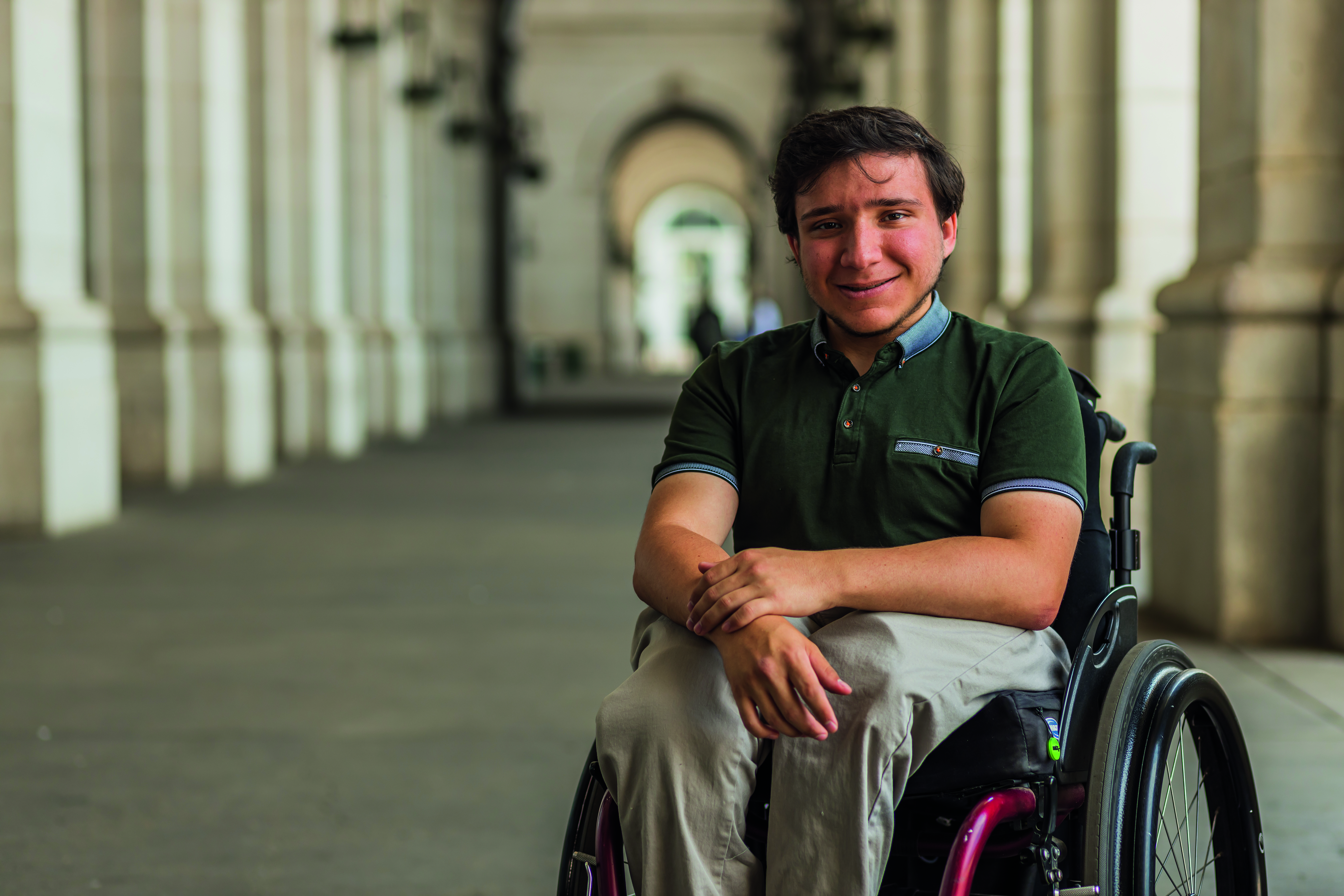 Person using a wheelchair sits in a long, arched stone colonnade, with soft natural light highlighting the architectural details of the walkway.