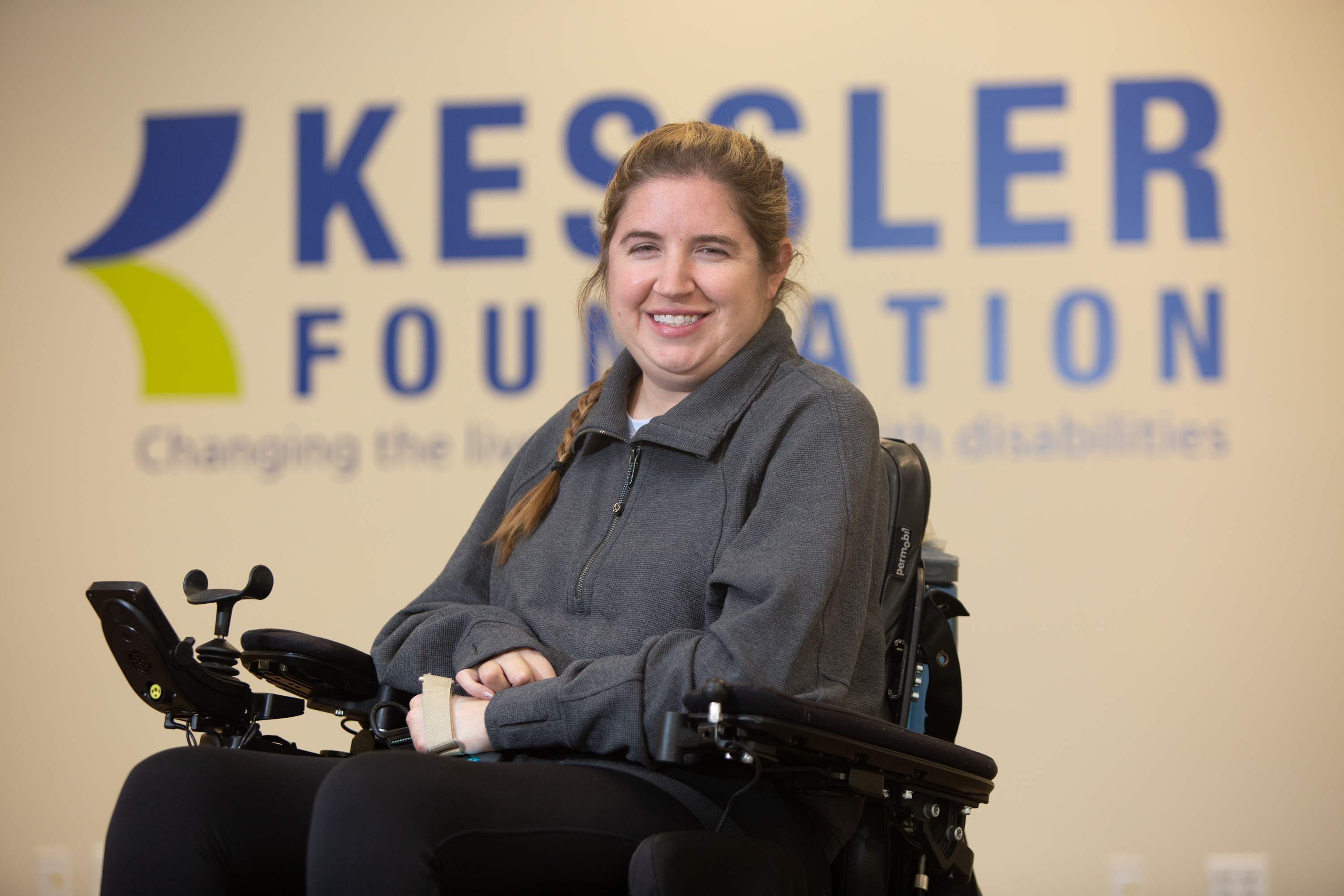A person seated in a power wheelchair in front of a Kessler Foundation backdrop