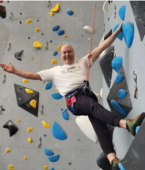Person wearing a “Up Ending Parkinson’s” T-shirt climbs an indoor rock wall, secured by a rope and harness, stretching one arm outward while balancing on blue and yellow holds against a gray wall.