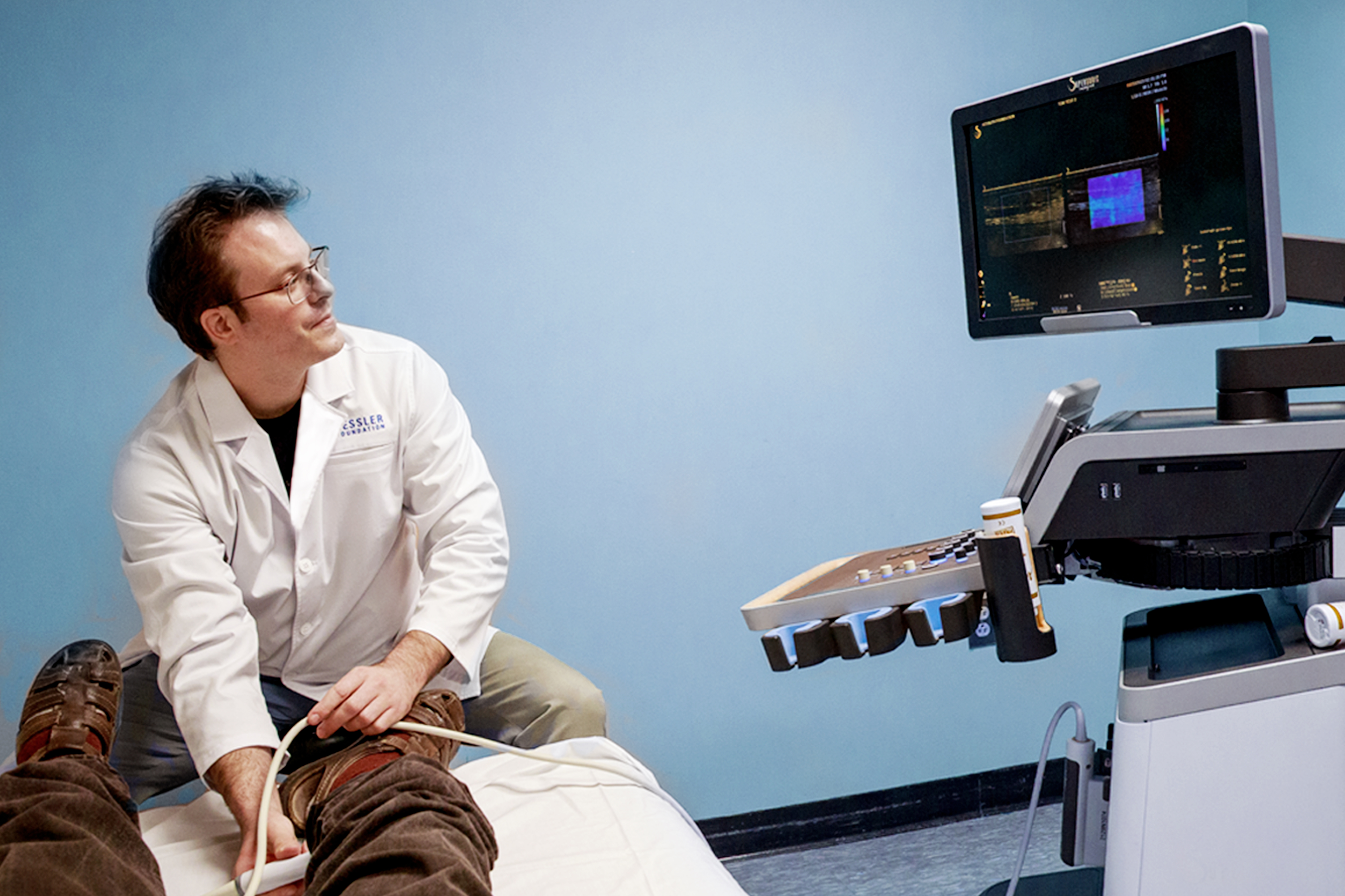 A patient participating in a regenerative rehabilitation session in a clinical setting, seated on an exam table with medical monitoring equipment positioned nearby as part of therapy and assessment.