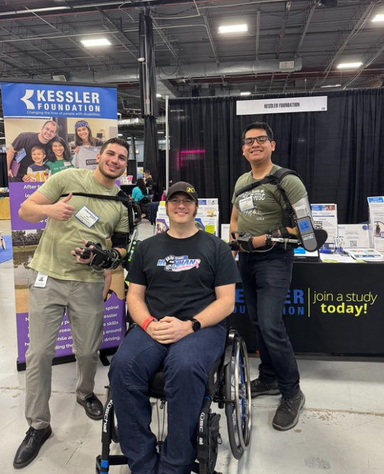 Three adults stand behind a participant using a wheelchair at a Kessler Foundation exhibit booth, with two individuals wearing robotic exoskeleton devices, demonstrating rehabilitation technology in a conference setting.