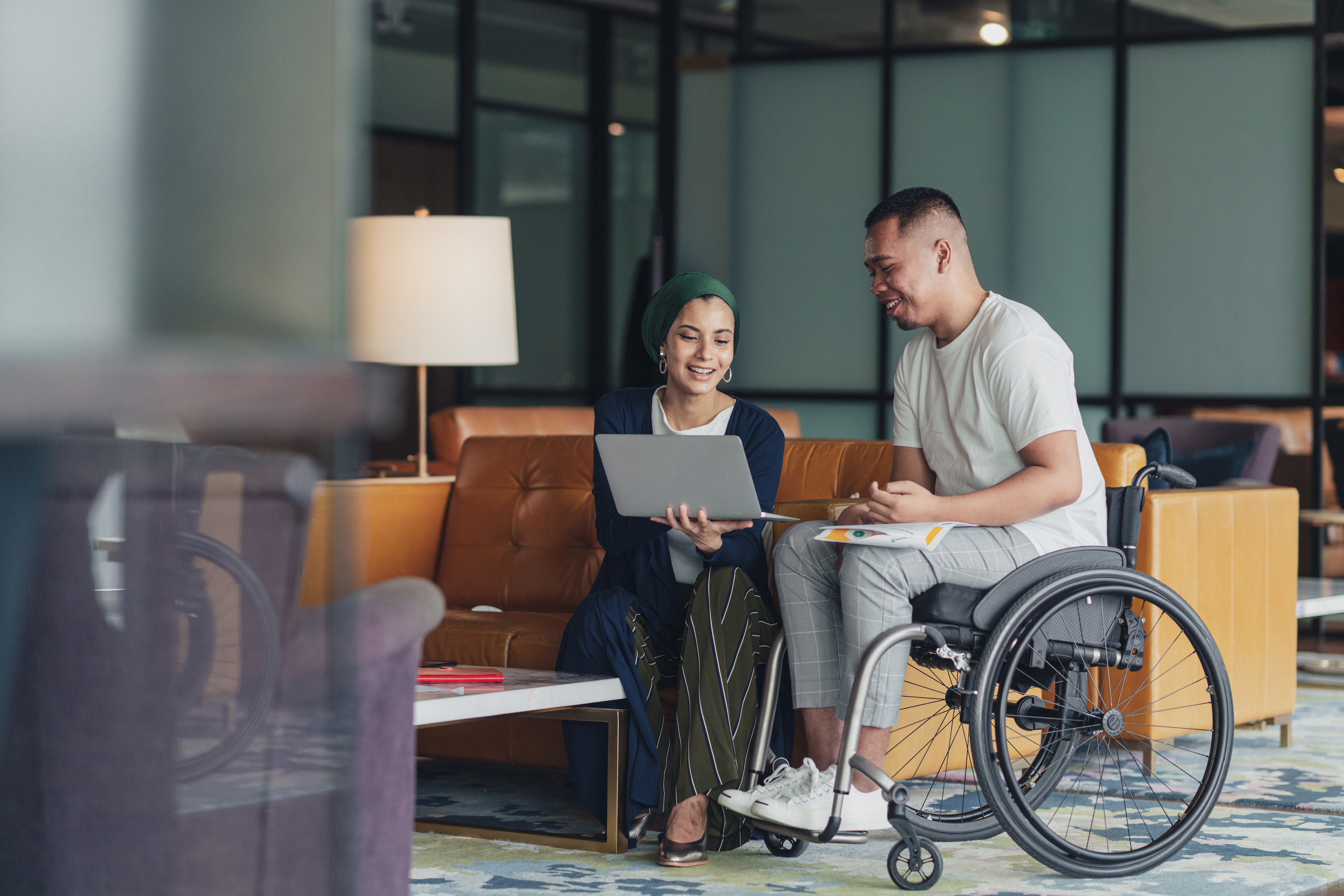 Small group meeting in a modern office lounge, with one participant using a wheelchair while reviewing notes on a laptop.