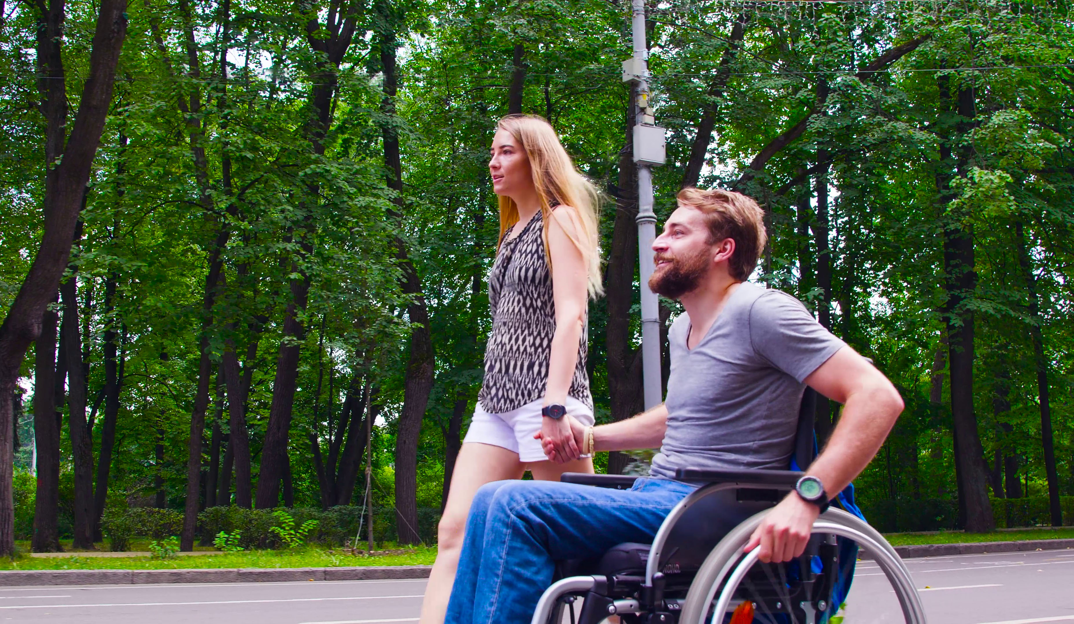 A couple holds hands while on a stroll in nature. The man is using a wheelchair.