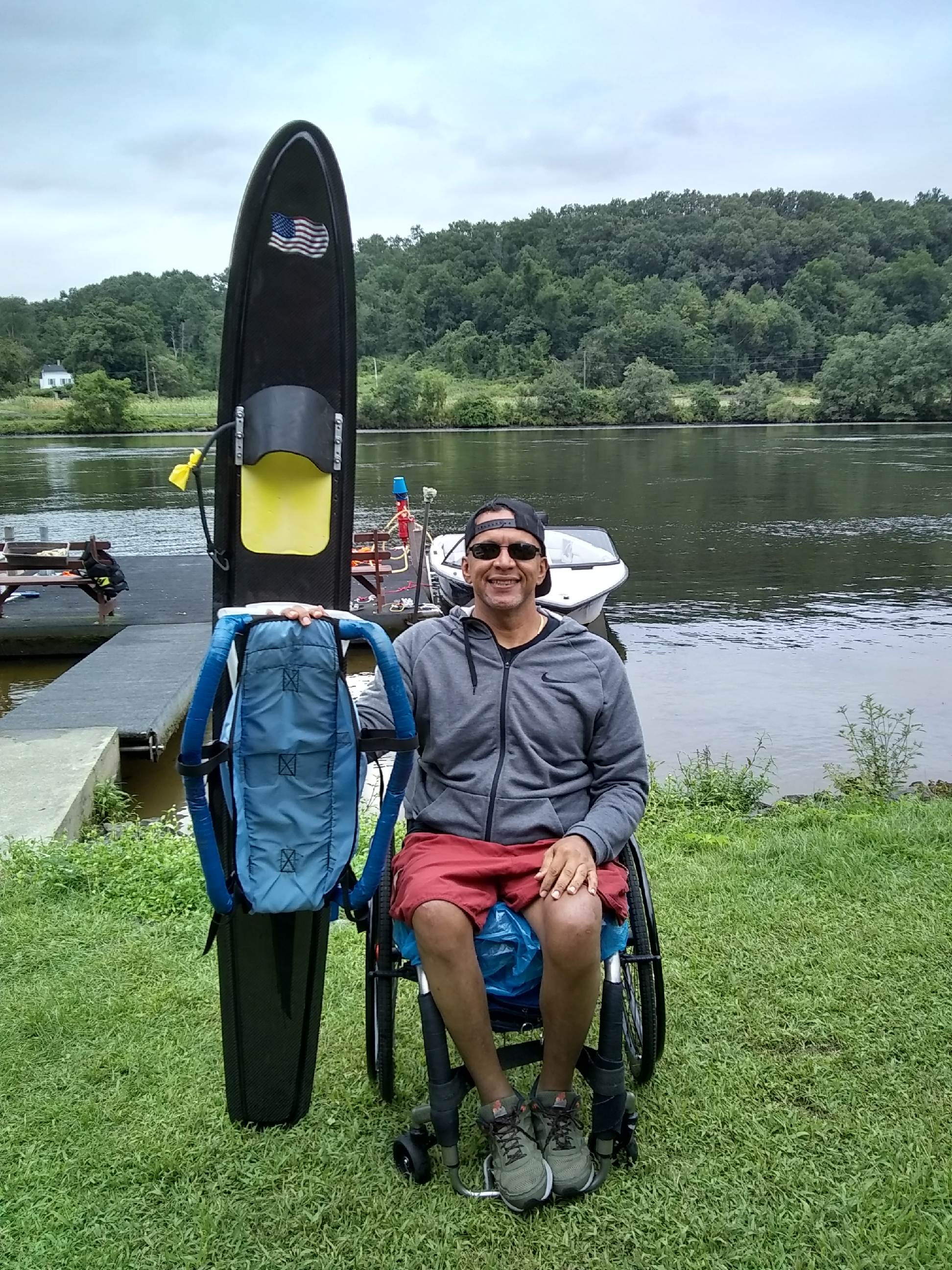A wheelchair user sits on grass near a lake, posing beside upright  outdoor gear, with calm water, boats, and a green shoreline in the background.