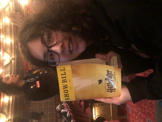 A person seated in an ornate theater holds a yellow showbill for Harry Potter and the Cursed Child, with rows of red seats, decorative balconies, and other audience members visible in the warmly lit auditorium.