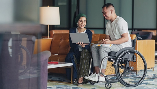 Small group meeting in a modern office lounge, with one participant using a wheelchair while reviewing notes on a laptop.