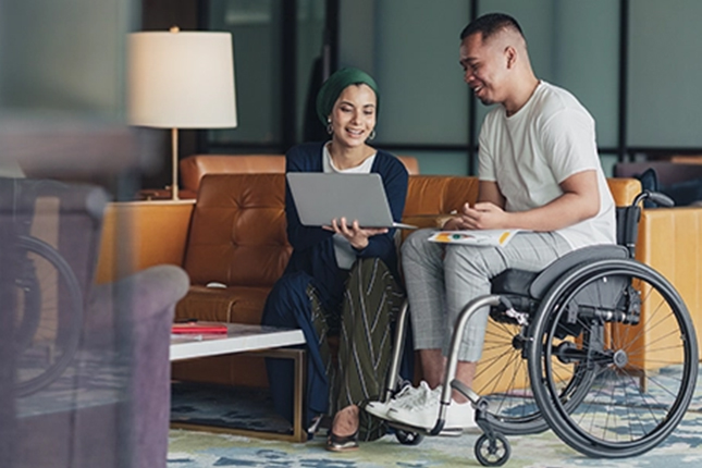 Small group meeting in a modern office lounge, with one participant using a wheelchair while reviewing notes on a laptop.