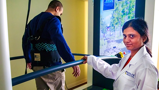 Kiran Karunakaran, PhD wearing a white lab coat standing beside a treadmill, monitoring another individual walking with a safety harness and support bars, in a rehabilitation setting with a virtual reality screen in the background.
