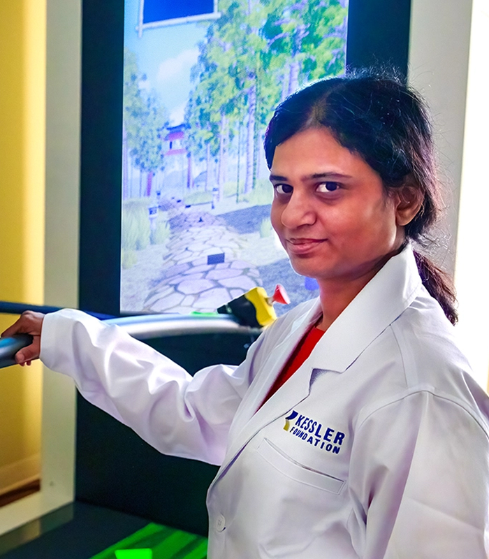 Kiran Karunakaran, PhD wearing a white lab coat standing beside a treadmill, monitoring another individual walking with a safety harness and support bars, in a rehabilitation setting with a virtual reality screen in the background.