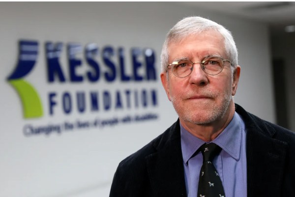 Gray-haired man with glasses, a blue shirt, dark blazer and tie, is pictured in front of a "Kessler Foundation" sign.