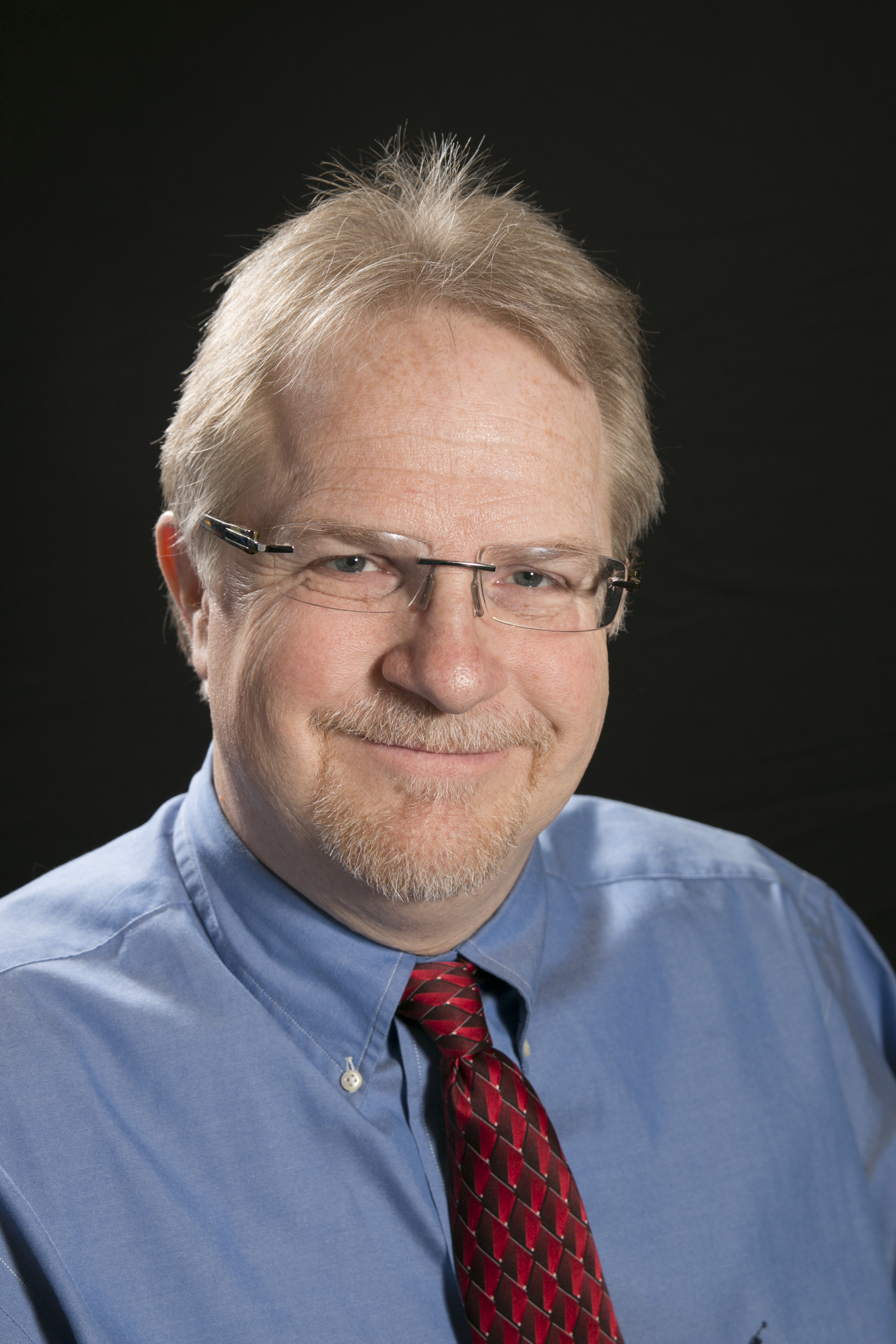 Image of Dr. Dyson-Hudson. He has light hair and glasses, wearing a blue button-up shirt and a red patterned tie.
