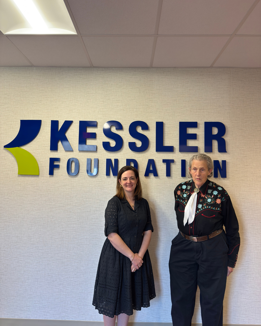 image of Helen Genova, PhD, and Temple Grandin, PhD, posing in front of the Kessler Foundation logo
