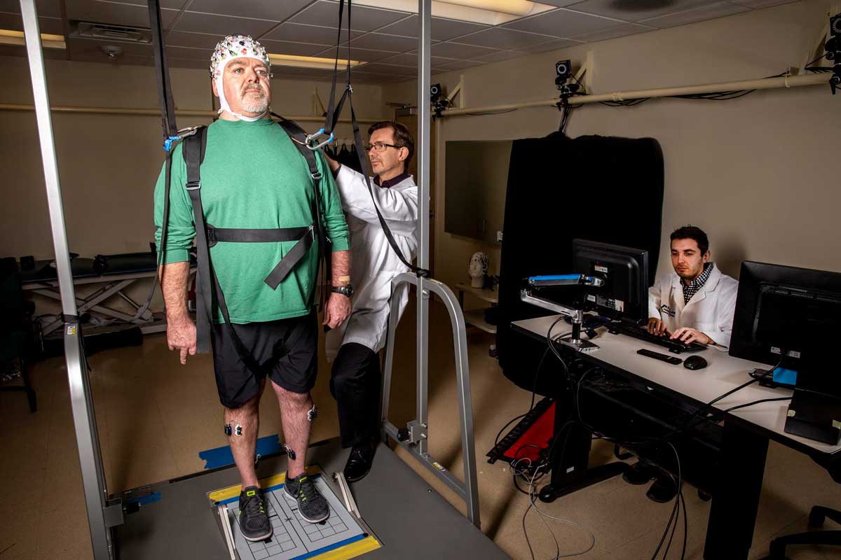 A study session being conducted in Dr Allexandre’s laboratory at Kessler Foundation.
Caption: A participant on the computerized balance platform wearing a specialized EEG device is prepared for the study by Dr. Allexandre.
