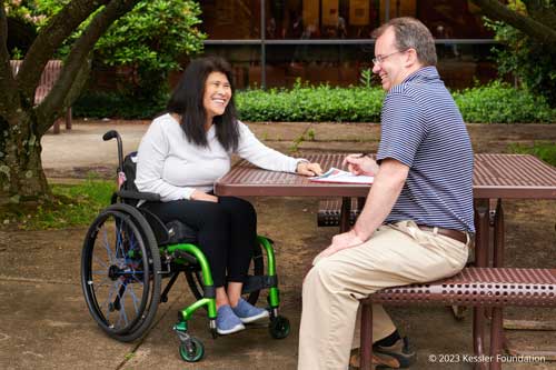 A woman in a wheelchair is sitting at a picnic table next to a man. They are both smiling.