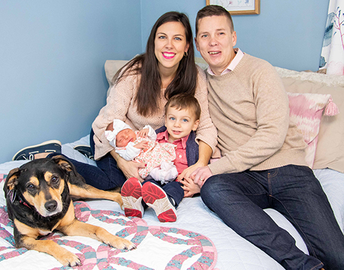 Ali and Pete Welch sitting on a bed with their son, Patrick, newborn daughter, Katie, and their dog.