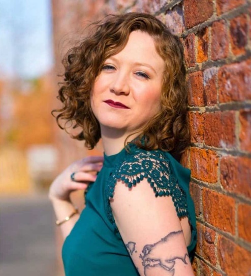 Image of Mimi Hayes leaning against a brick wall. She is wearing a green shirt with lace trim.