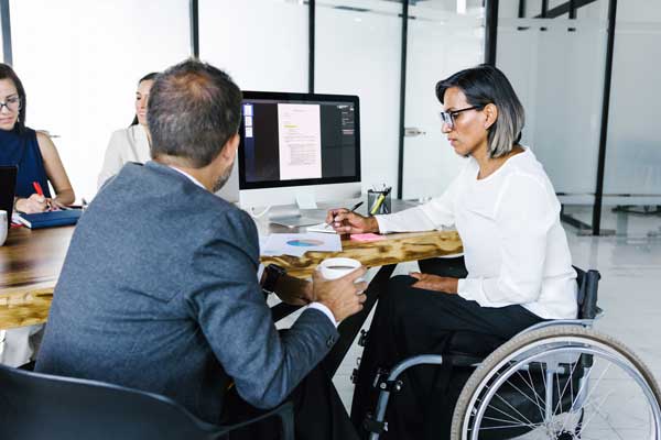 A man in a suit and a woman in a wheelchair are sitting at a desk in an office working together.