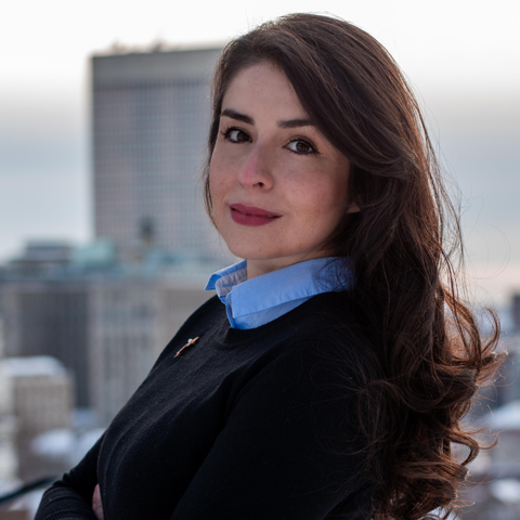Woman with a dark jacket, blue shirt, long brown hair at rooftop in a city