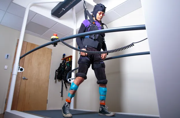 A person wearing a safety harness and motion-tracking headgear walks on a curved treadmill in a clinical rehabilitation lab, with padded knee supports, taped lower legs, and sneakers, while suspended for assisted gait training.