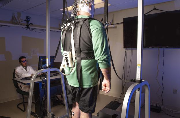 A study participant wearing a safety harness stands on a raised walking platform while undergoing balance training. The participant is fitted with a brain stimulation cap and motion sensors, as a researcher monitors the session from a computer in a laboratory setting.