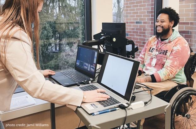 Research Assistant Caroline Armknecht sits in front of two laptops and across from a black male study participant in a wheelchair. She is administering a saccadic eye movement exercise using the Eyelink Portable Duo tracking system for a study involving individuals with spinal cord injury.