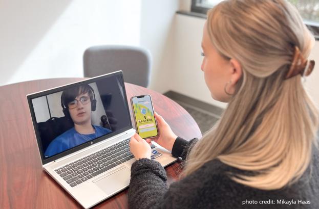 Center for Autism Research Course Developer Mikayla Haas sits at a table with a laptop displaying a study participant on the screen. She holds a smartphone with a visual of the KF-Stride app open.