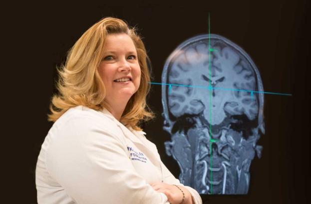 Person wearing a white lab coat standing in front of a large brain scan image displayed on a screen, with green crosshair lines marking sections of the brain.