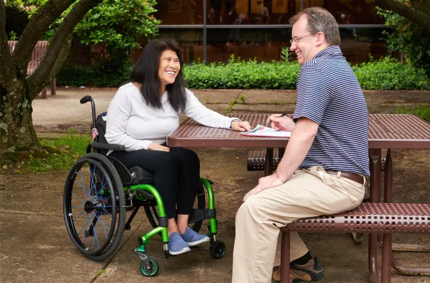 Accessible outdoor setting with a person in a wheelchair and another individual reviewing papers at a picnic table. 