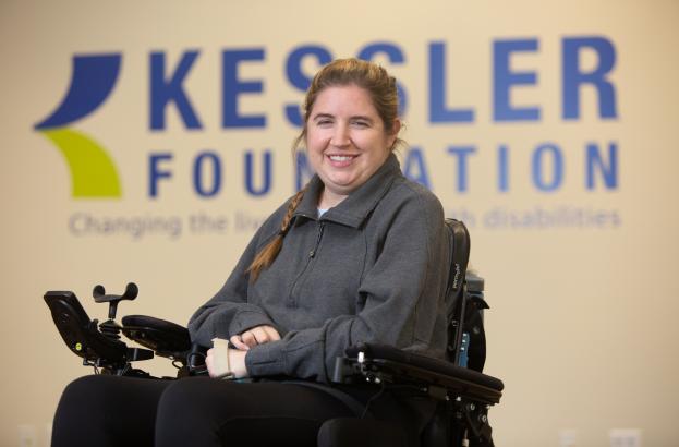 A person seated in a power wheelchair in front of a Kessler Foundation backdrop