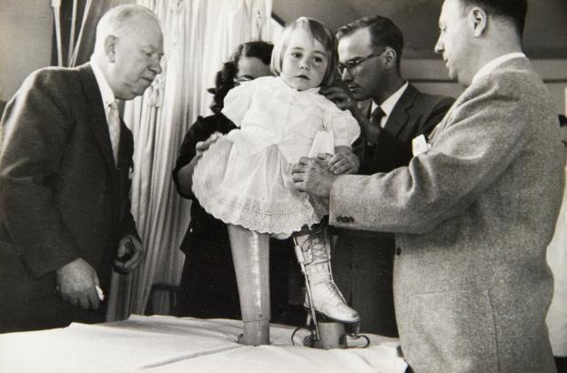Black-and-white photograph of a child supported by several adults during a rehabilitation session.