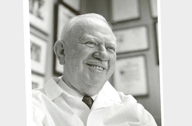 A man smiles in front of a wall of framed art, degrees, and certificates