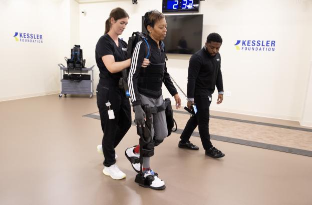 Two clinicians assist a research participant wearing a robotic exoskeleton device while walking along an indoor therapy path at Kessler Foundation, with monitoring equipment and a wall-mounted display visible in the background.