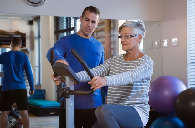A woman uses a stationary bike in a rehabilitation setting with a therapist providing guidance 