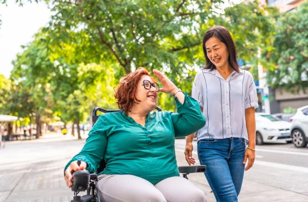 Two people on a sidewalk outdoors. One is using a powerwheel chair. The two are smiling and gesturing at one another.
