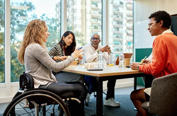 Small group of diverse individuals in a work setting. One individual is using a wheelchair and is seated at the head of the conference table
