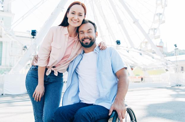 a person using a wheelchair and their caregiver smile together in front of a Ferris wheel